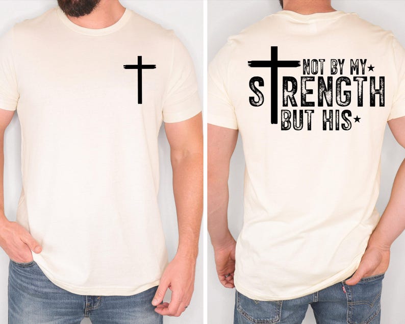 May include: Cream-colored t-shirt with a black cross on the front and back. The back of the shirt features the text "NOT BY MY STRENGTH BUT HIS" in a distressed font. The shirt is a short-sleeved crew neck.