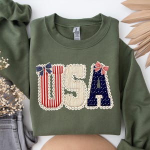 May include: An olive green sweatshirt featuring the word "USA" in a decorative applique. The letters are adorned with red and white stripes, blue stars, and a cream-coloured textured fabric, edged with lace. Two decorative bows are attached.