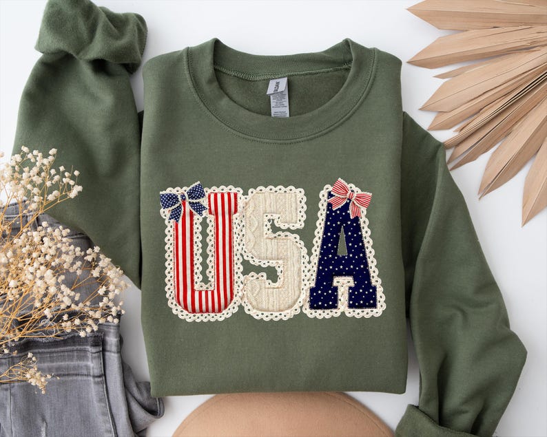 May include: An olive green sweatshirt featuring the word "USA" in a decorative applique. The letters are adorned with red and white stripes, blue stars, and a cream-coloured textured fabric, edged with lace. Two decorative bows are attached.