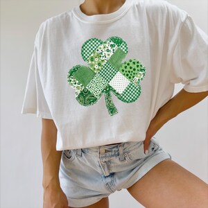 May include: White t-shirt featuring a patchwork shamrock design in various shades of green. The shamrock is composed of different patterns, including floral, gingham, and paisley. The shirt is a casual, short-sleeved style.