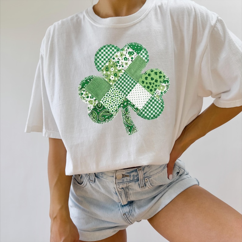 May include: White t-shirt featuring a patchwork shamrock design in various shades of green. The shamrock is composed of different patterns, including floral, gingham, and paisley. The shirt is a casual, short-sleeved style.