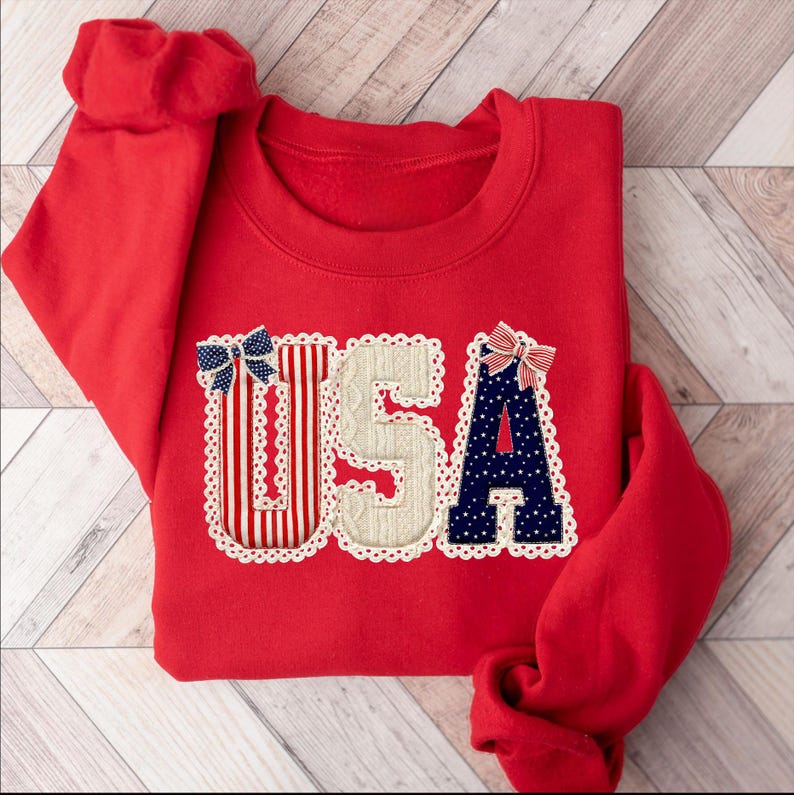 May include: A red sweatshirt featuring the word "USA". The letters are decorated with red and white stripes, a cream-coloured knit pattern, and navy blue with white stars. Each letter is outlined with white lace and topped with a bow.
