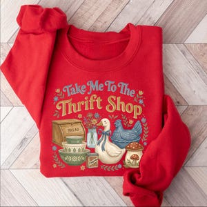 May include: A red sweatshirt with the phrase "Take Me To The Thrift Shop" in a vintage font. The graphic includes a bread box, bowls, a duck with a blue bow, a blue chicken, and a mushroom. Floral accents surround the design.
