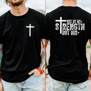 May include: Black t-shirt with a white cross on the front. The back of the shirt features a white cross and the text "NOT BY MY STRENGTH BUT HIS". The shirt is made of a soft material.