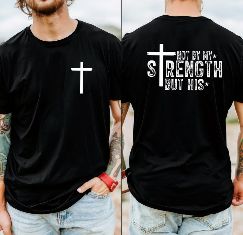 May include: Black t-shirt with a white cross on the front. The back of the shirt features a white cross and the text "NOT BY MY STRENGTH BUT HIS". The shirt is made of a soft material.
