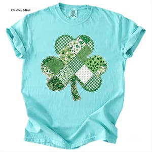 May include: A light blue t-shirt featuring a large shamrock design. The shamrock is made of various green fabric patterns, including floral, plaid, and paisley. The text "Chalky Mint" is in the upper left corner of the image.