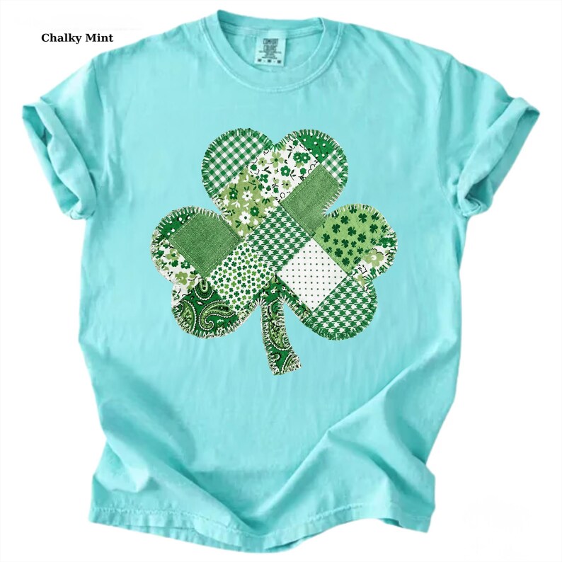 May include: A light blue t-shirt featuring a large shamrock design. The shamrock is made of various green fabric patterns, including floral, plaid, and paisley. The text "Chalky Mint" is in the upper left corner of the image.