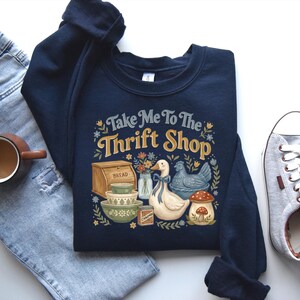 May include: Navy blue sweatshirt with the text "Take Me To The Thrift Shop" and a graphic of a duck, chicken, mushroom, bread, and bowls. The sweatshirt is paired with light blue jeans, a cup of coffee, and gray sneakers.