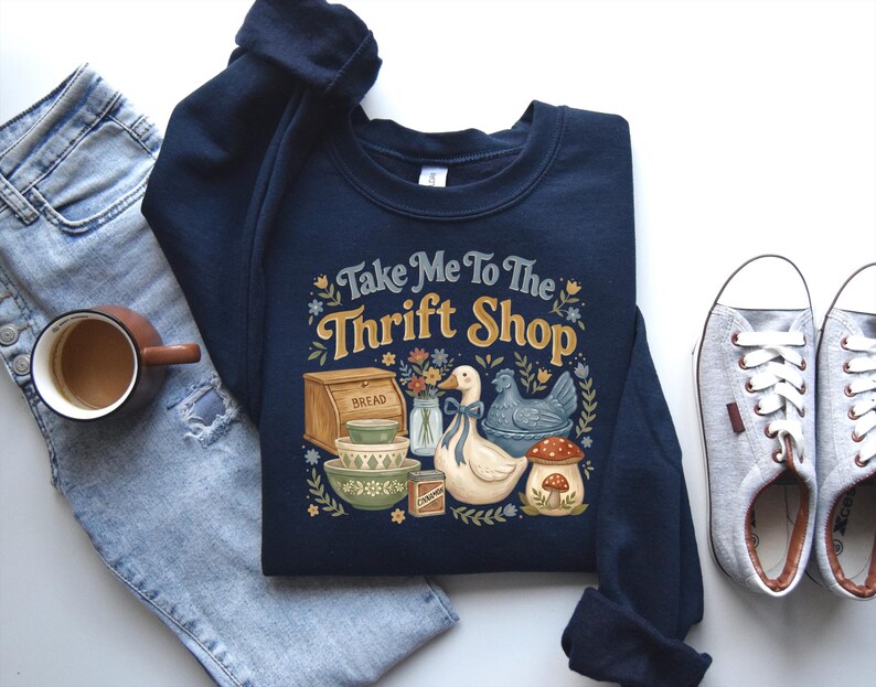 May include: Navy blue sweatshirt with the text "Take Me To The Thrift Shop" and a graphic of a duck, chicken, mushroom, bread, and bowls. The sweatshirt is paired with light blue jeans, a cup of coffee, and gray sneakers.