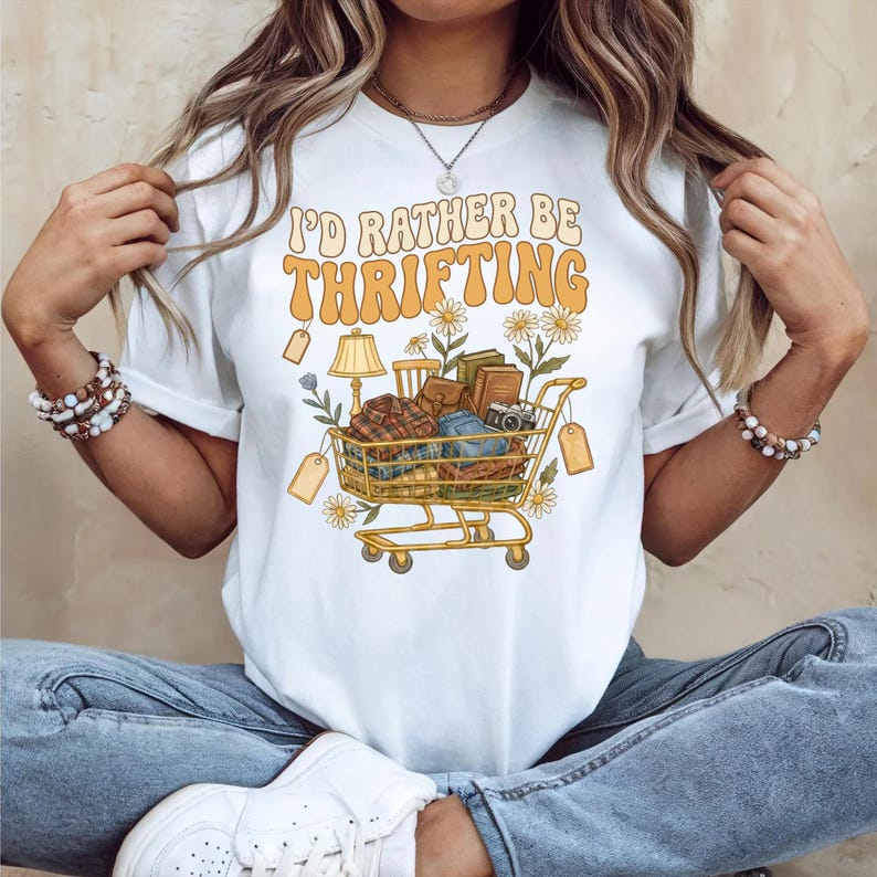Op de afbeelding: Wit T-shirt met de tekst "I'D RATHER BE THRIFTING" in oranje en gele retro letters. Een winkelwagenafbeelding is gevuld met vintage items zoals boeken, een lamp en kleding. Madeliefjes en prijskaartjes omringen de kar.