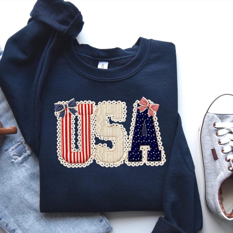 Three large letters spelling U S A arranged with printed textures simulating knitwear, starry fabric, and striped material. The typography is bordered by a printed lace edge and decorated with faux fabric bows.