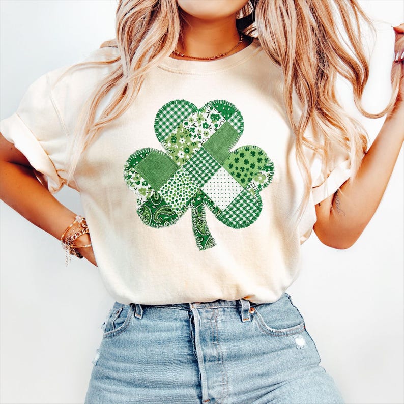May include: Cream-colored t-shirt featuring a large, patchwork shamrock design. The shamrock is composed of various green patterns, including gingham, floral, and paisley. The shirt is paired with blue denim jeans, creating a casual, everyday look.