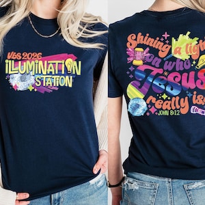 May include: Navy blue t-shirt with the text "VBS 2026 ILLUMINATION STATION" on the front. The back of the shirt has the text "Shining a light on who Jesus really is!" with "John 8:12". The design includes colorful graphics.