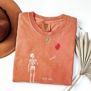 Pode incluir: Uma t-shirt cor de terracota com um gr&aacute;fico branco de um esqueleto a olhar para um bal&atilde;o vermelho segurado por uma m&atilde;o esquel&eacute;tica destacada. O texto "Well, shit..." est&aacute; impresso a branco por baixo do gr&aacute;fico. A t-shirt tem gola redonda e uma etiqueta Comfort Colors.