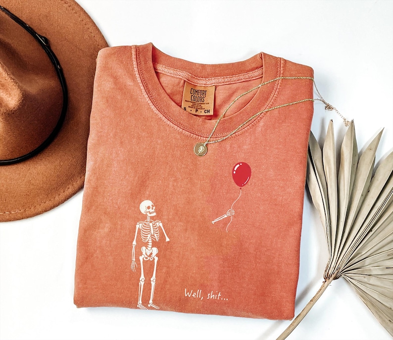 Pode incluir: Uma t-shirt cor de terracota com um gr&aacute;fico branco de um esqueleto a olhar para um bal&atilde;o vermelho segurado por uma m&atilde;o esquel&eacute;tica destacada. O texto "Well, shit..." est&aacute; impresso a branco por baixo do gr&aacute;fico. A t-shirt tem gola redonda e uma etiqueta Comfort Colors.