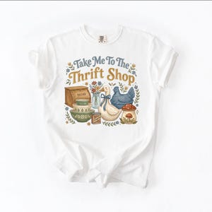 May include: White t-shirt with a colorful graphic that reads "Take Me To The Thrift Shop." The design features a bread box, bowls, a duck, a chicken, a mushroom, and other vintage-style elements.