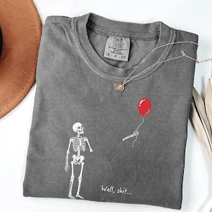 Pode incluir: Uma t-shirt de manga curta cinzento-carv&atilde;o com um gr&aacute;fico branco de um esqueleto a olhar para uma m&atilde;o esquel&eacute;tica separada que segura um bal&atilde;o vermelho. O texto "Well, shit..." est&aacute; impresso a branco por baixo do gr&aacute;fico. Gola redonda.
