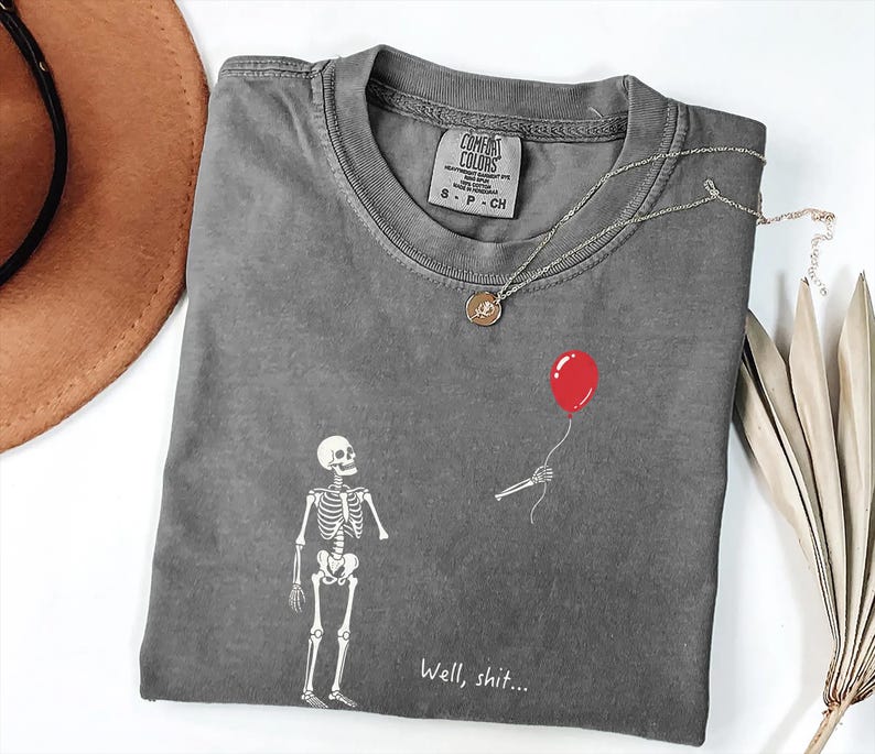 Pode incluir: Uma t-shirt de manga curta cinzento-carv&atilde;o com um gr&aacute;fico branco de um esqueleto a olhar para uma m&atilde;o esquel&eacute;tica separada que segura um bal&atilde;o vermelho. O texto "Well, shit..." est&aacute; impresso a branco por baixo do gr&aacute;fico. Gola redonda.