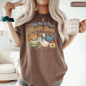 May include: Espresso-colored t-shirt with the text "Take Me To The Thrift Shop" and a graphic of a bread box, bowls, a duck, a chicken, and a mushroom. The shirt is a comfortable, casual style.