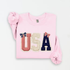 May include: A pale pink sweatshirt featuring the letters "USA". The "U" is red and white striped, the "S" is beige, and the "A" is navy blue with white dots. Each letter is adorned with a decorative bow, in patriotic colours.