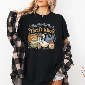 May include: Black t-shirt with the text "Take Me To The Thrift Shop" in yellow and blue. The graphic includes illustrations of a bread box, bowls, a duck, a chicken, and a mushroom.