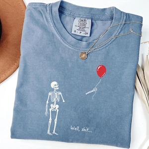 Pode incluir: Uma t-shirt azul de manga curta com um gr&aacute;fico branco de um esqueleto a olhar para um bal&atilde;o vermelho flutuante segurado por uma m&atilde;o esquel&eacute;tica desencarnada. O texto "Well, shit..." est&aacute; impresso a branco sob o esqueleto. Etiqueta Comfort Colors.