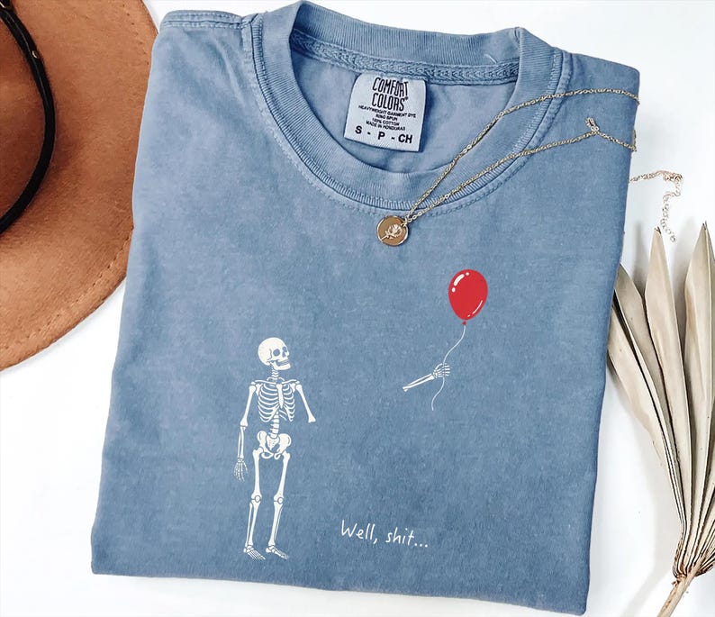 Pode incluir: Uma t-shirt azul de manga curta com um gr&aacute;fico branco de um esqueleto a olhar para um bal&atilde;o vermelho flutuante segurado por uma m&atilde;o esquel&eacute;tica desencarnada. O texto "Well, shit..." est&aacute; impresso a branco sob o esqueleto. Etiqueta Comfort Colors.