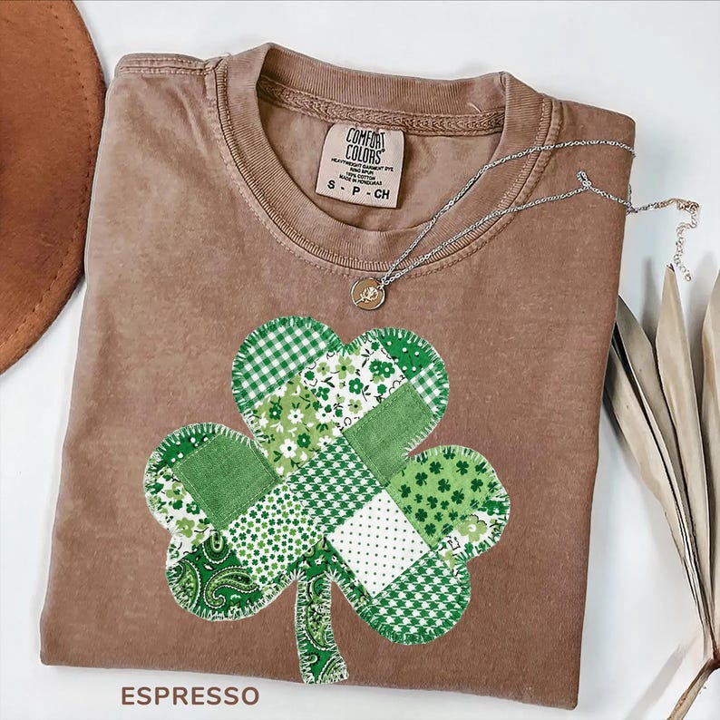 May include: Espresso-colored t-shirt with a patchwork shamrock design in various shades of green. A silver necklace with a small pendant rests on the shirt. The shirt is labeled "Comfort Colors" and is size small.