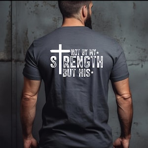 May include: Dark gray t-shirt with a white cross and the text "NOT BY MY STRENGTH BUT HIS". The shirt has short sleeves and a crew neck. The text is in a bold, distressed font.