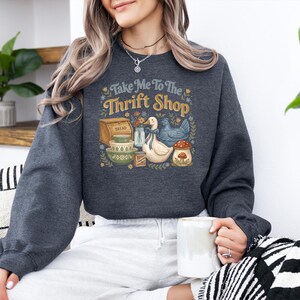 May include: Dark gray sweatshirt with a colorful graphic that reads "Take Me To The Thrift Shop." The graphic features a bread box, bowls, a duck, a chicken, and a mushroom. The sweatshirt is made of a soft material.