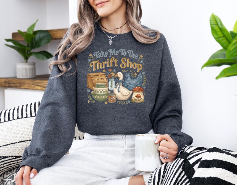 May include: Dark gray sweatshirt with a colorful graphic that reads "Take Me To The Thrift Shop." The graphic features a bread box, bowls, a duck, a chicken, and a mushroom. The sweatshirt is made of a soft material.