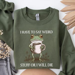 May include: Olive green sweatshirt with the text "I HAVE TO SAY WEIRD STUFF OR I WILL DIE." A cartoon frog holding a coffee mug is printed below the text. The sweatshirt is a crew neck style.