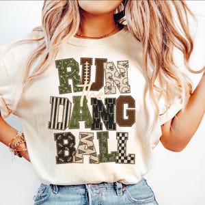 May include: A cream-colored t-shirt featuring the text RUN THE DANG BALL in large, block-style letters. Each letter is filled with various football-themed patterns in shades of olive green, black, brown, and tan, including helmets and balls.