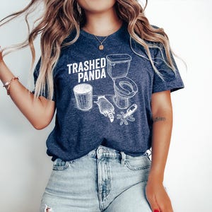 May include: A navy blue t-shirt with the text "Trashed Panda" and a white graphic of a toilet, trash can, raccoon, and a spilled bottle. The shirt is made of a soft material and has short sleeves.