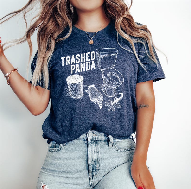 May include: A navy blue t-shirt with the text "Trashed Panda" and a white graphic of a toilet, trash can, raccoon, and a spilled bottle. The shirt is made of a soft material and has short sleeves.