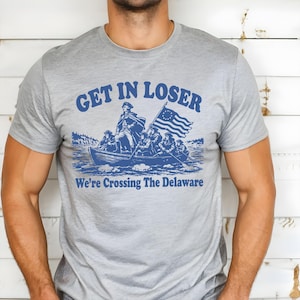 May include: A gray t-shirt with a blue graphic of a boat crossing a river, featuring a flag and the text "GET IN LOSER" above "We're Crossing The Delaware". The design is a parody of the famous painting.
