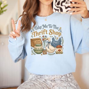 May include: Light blue sweatshirt with the text "Take Me To The Thrift Shop" and a graphic of a bread box, bowls, a duck, a chicken, and a mushroom. The sweatshirt is a soft, comfortable material.
