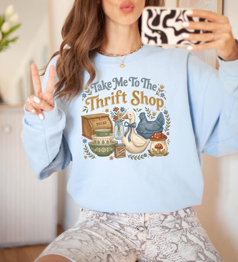 May include: Light blue sweatshirt with the text "Take Me To The Thrift Shop" and a graphic of a bread box, bowls, a duck, a chicken, and a mushroom. The sweatshirt is a soft, comfortable material.