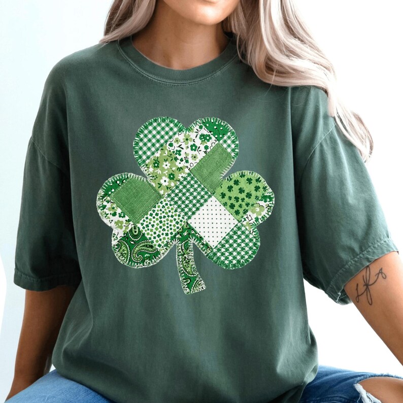 Detailed view of a four-leaf clover motif composed of various green patterned squares. The patchwork includes traditional paisley, plaid, and clover prints connected by visible white thread stitching. The design sits centered on a high-quality garment.