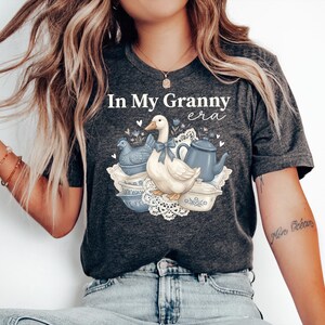May include: Dark gray t-shirt with the text "In My Granny era" above a graphic of a white goose with a blue bow, a blue teapot, and blue dishes. The shirt is made of a soft material.