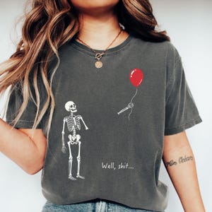 May include: A charcoal gray t-shirt featuring a white skeleton graphic. The skeleton is missing an arm, which is shown floating separately while holding a single red balloon. The text "Well, shit..." is printed in white below the skeleton.