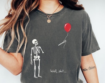 Peut inclure: Un t-shirt gris anthracite avec un motif de squelette blanc. Le squelette a un bras détaché qui flotte à côté, tenant un ballon rouge. Le texte "Well, shit..." est imprimé en blanc sous le squelette sur le devant du vêtement.