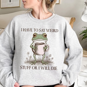 May include: Light gray sweatshirt with the text "I HAVE TO SAY WEIRD STUFF OR I WILL DIE." The graphic features a cartoon frog holding a coffee mug. The frog is green with large eyes and is standing in grass.