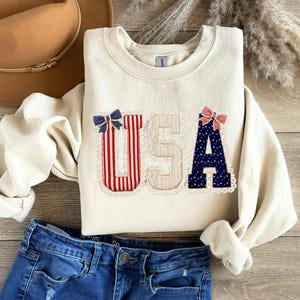 May include: Cream-coloured sweatshirt with the word "USA" appliqu&eacute;d on the front. The letters are decorated with red and white stripes, blue polka dots, and bows. A brown hat and blue jeans are also in the image.