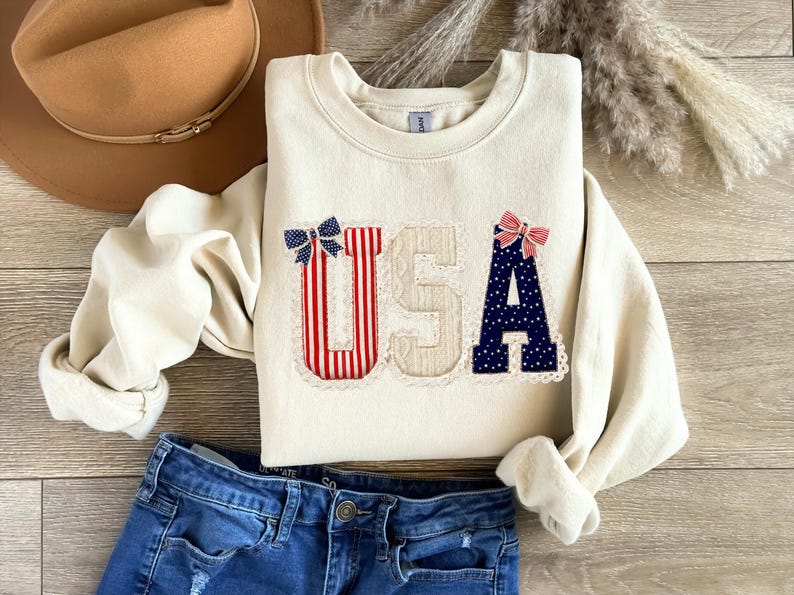 May include: Cream-coloured sweatshirt with the word "USA" appliqu&eacute;d on the front. The letters are decorated with red and white stripes, blue polka dots, and bows. A brown hat and blue jeans are also in the image.