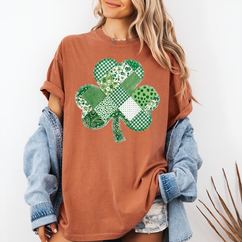May include: A rust-colored t-shirt featuring a large, patchwork shamrock design in various shades of green and white. The shamrock is centered on the shirt, with a detailed, textured appearance. The shirt has rolled-up sleeves and is paired with a denim jacket.