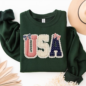 May include: A dark green sweatshirt featuring the letters "USA" appliqu&eacute;d on the front. The letters are decorated with red, white, and blue patterns and bows, and trimmed with white lace. The sleeves have dark green cuffs.