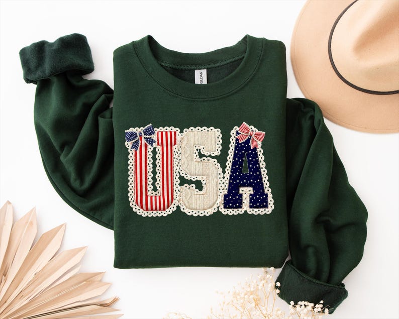 May include: A dark green sweatshirt featuring the letters "USA" appliqu&eacute;d on the front. The letters are decorated with red, white, and blue patterns and bows, and trimmed with white lace. The sleeves have dark green cuffs.