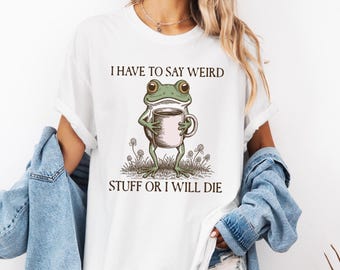 May include: White t-shirt featuring a cartoon frog holding a coffee mug. The frog is green with large eyes and is standing in a field of flowers. The text on the shirt reads "I HAVE TO SAY WEIRD STUFF OR I WILL DIE."