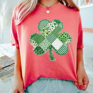May include: A coral-colored t-shirt featuring a large, patchwork shamrock design. The shamrock is composed of various green patterned fabrics, including gingham, floral, and paisley prints. The shirt has short sleeves and a crew neck, perfect for St. Patrick's Day.